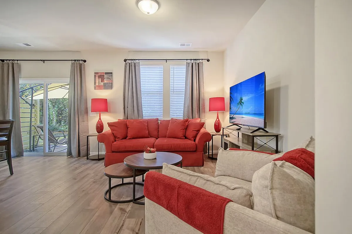 The image showcases a welcoming living room with a vibrant red sofa as the centerpiece, complemented by a coffee table, a television for entertainment, and natural light streaming through curtained windows. The cozy arrangement invites guests to relax, socialize, or enjoy quiet moments, making it ideal for family gatherings or unwinding after exploring Charleston.