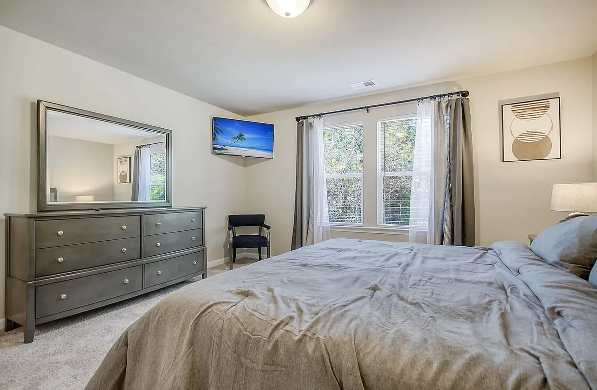 This image depicts a bright, serene bedroom featuring a comfortable bed, a dresser, windows allowing in natural light, and a television. The decor is inviting and restful, suggesting a peaceful retreat ideal for relaxation or ending the day with a favorite show.