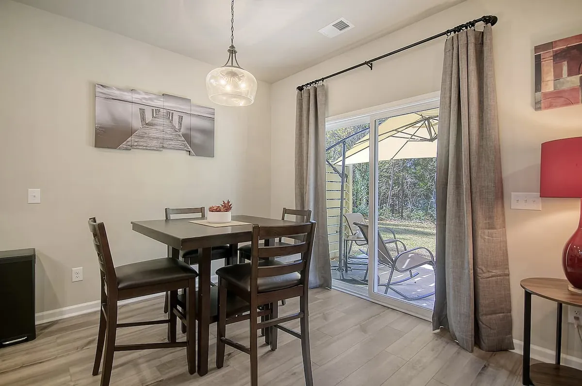 A well-lit dining area with a large table, hanging light fixture, sliding glass doors that let in natural light, and decorative wall art. The setup offers an inviting space for working, relaxing, and group gatherings, with comfortable seating that supports both productivity and downtime.