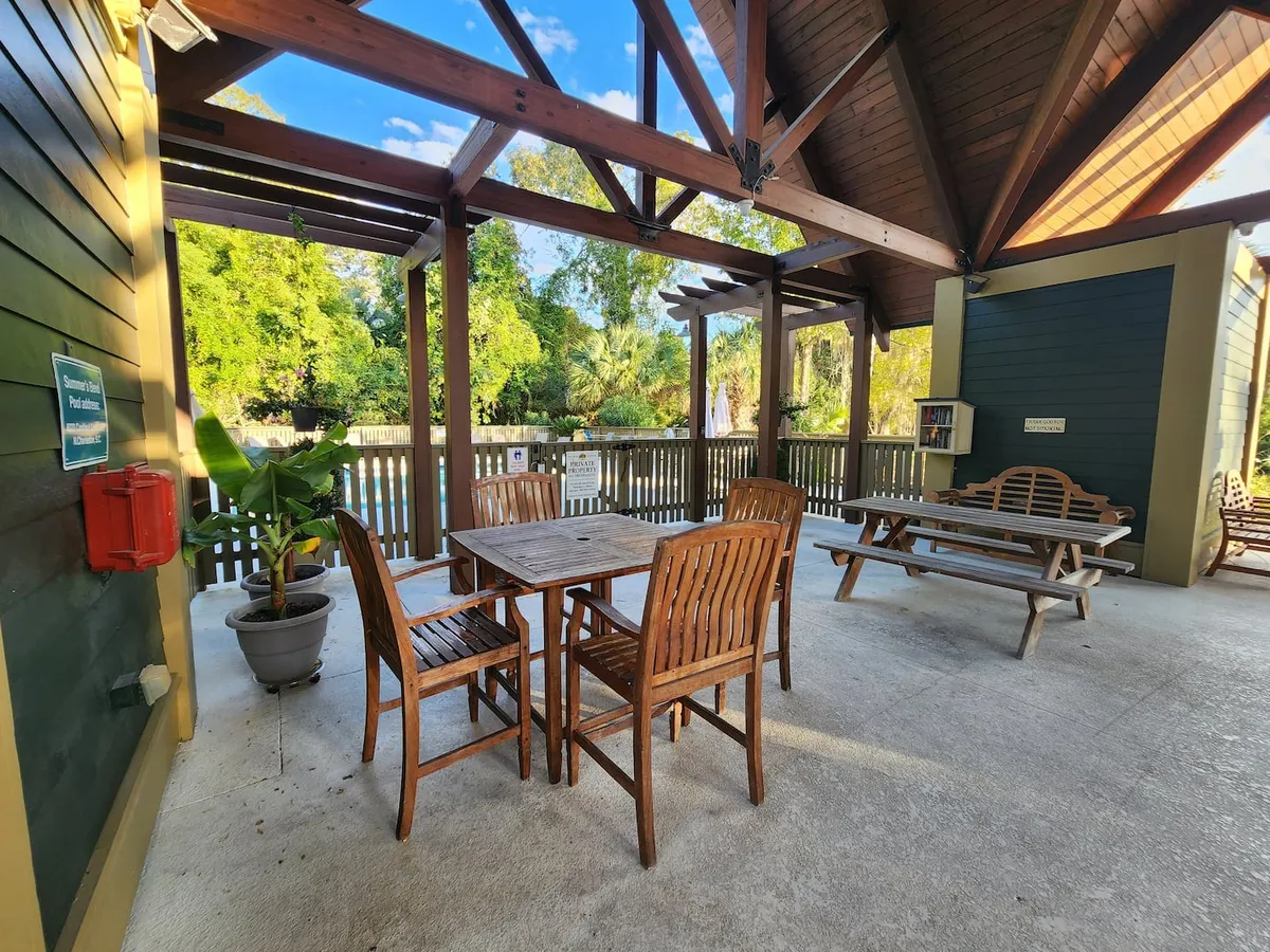 A wooden picnic table and dining chairs sit under a pergola, surrounded by lush greenery. The relaxed outdoor dining setup suggests moments of togetherness and joy, perfect for family meals or casual gatherings after a day of adventure. Sunlight filters through, creating a warm, inviting space ideal for connecting with loved ones.