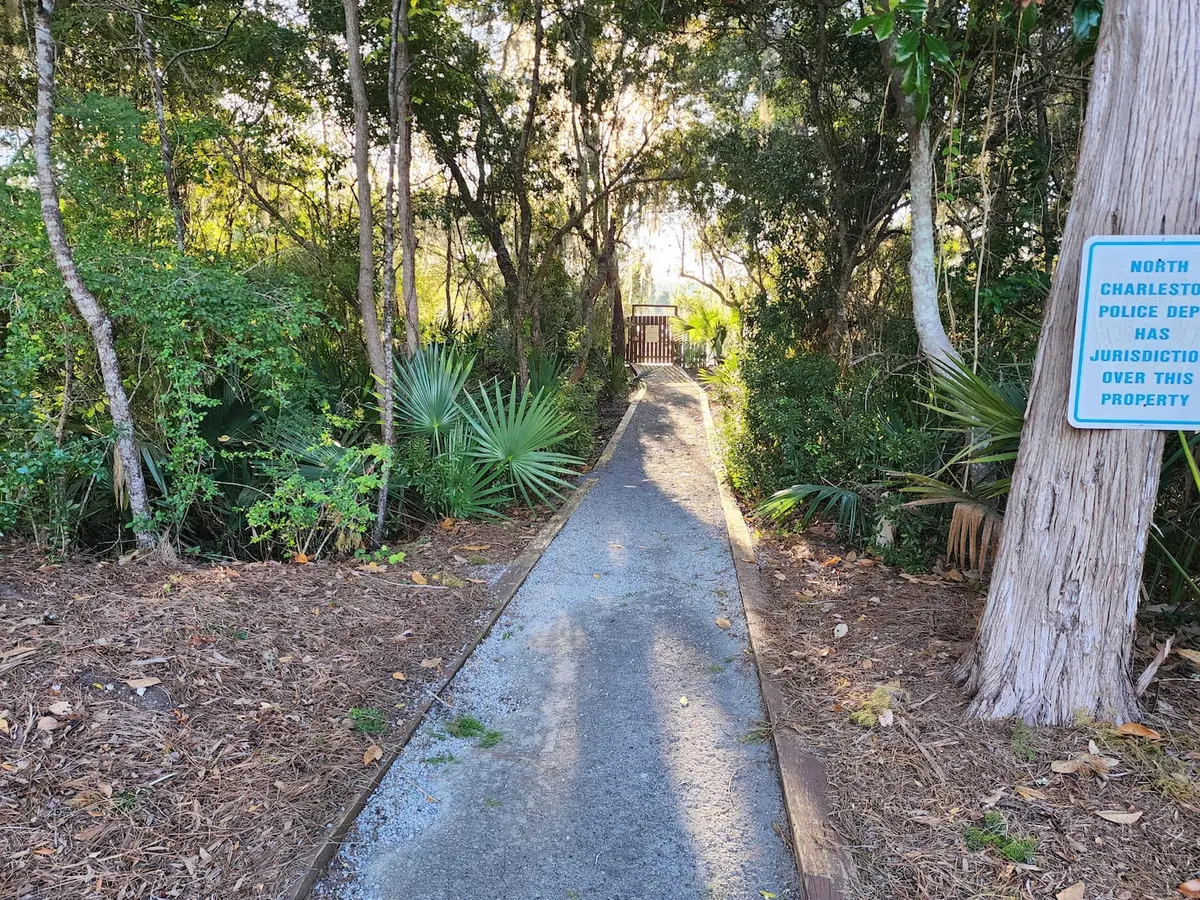 A quiet neighborhood pathway lined with mature trees, creating a serene, green environment. The scene includes a police jurisdiction sign, emphasizing a safe and peaceful atmosphere, and invites leisurely walks and nature exploration.