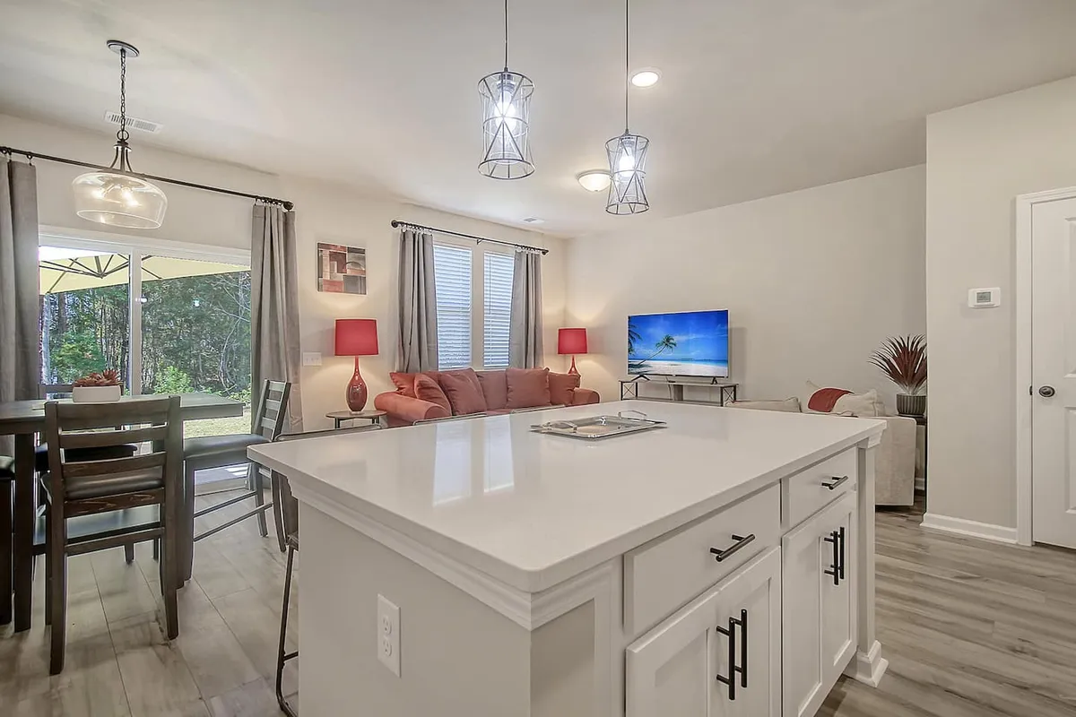 A spacious, sunlit open-concept main area featuring a modern kitchen island, a cozy dining space, and an inviting living area. Large windows flood the room with natural light, while comfortable seating and a flowing layout encourage gathering and conversation.