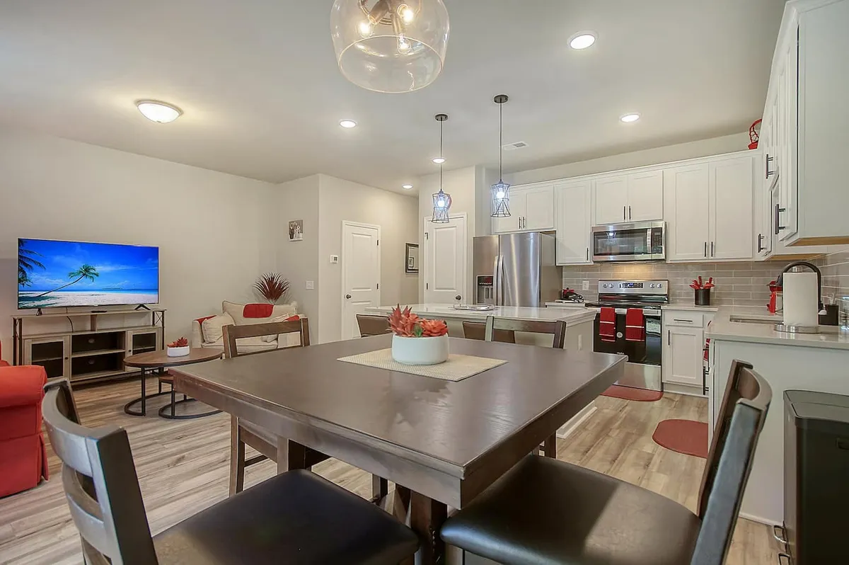 A welcoming dining room scene with a prominent dining table, kitchen appliances in the background, and a television. The setup is inviting for family meals or entertaining guests, and the background kitchen elements hint at the space’s functionality for group cooking or casual dining.